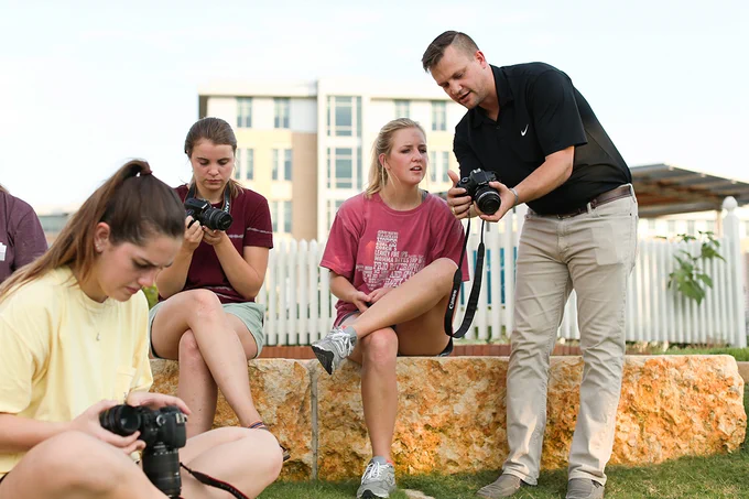 Man holds camera in front of student with other students sitting nearby. 
