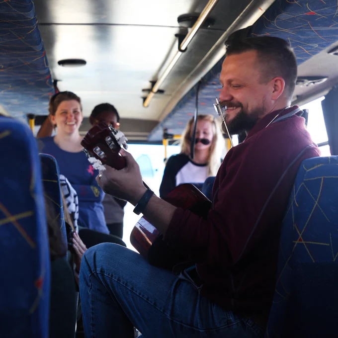 A man plays a guitar on a bus while students smile in the background. 