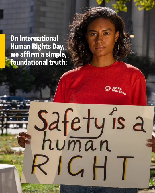 A woman wearing a red Safe Horizon t-shirt holds a hand-painted sign that reads "Safety is a human RIGHT" while standing outdoors in front of a large building. 