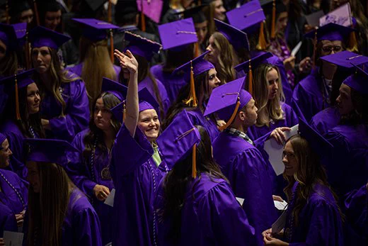 In a sea of Weber State graduates all wearing purple robes, a young woman raises her hand to wave at someone in the audience.