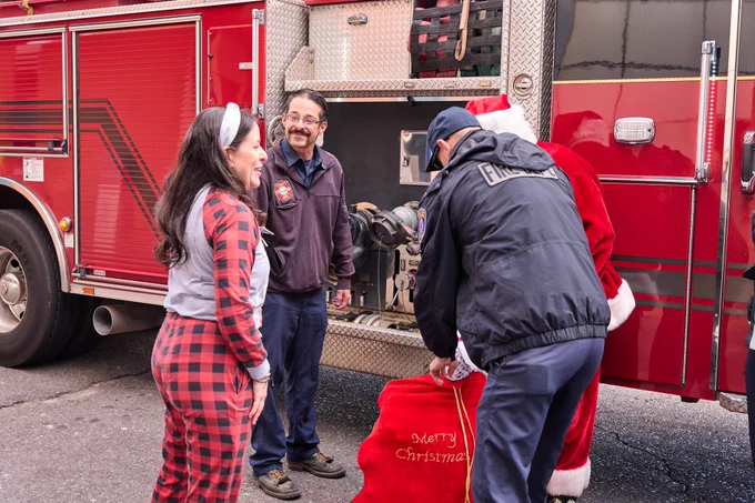Three adults interacting happily near a fire truck; one person is handing over a 'Merry Christmas' sack to another, with a firefighter observing nearby.