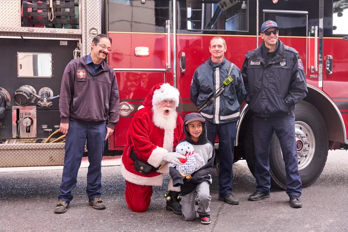 Santa Claus and a child are posing in front of a fire truck, accompanied by three firefighters.