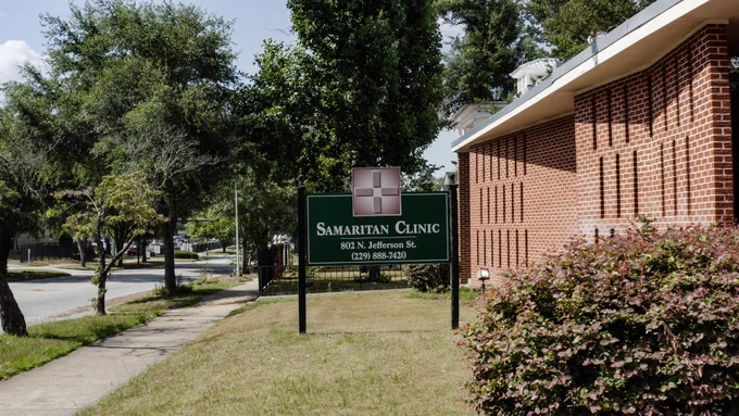 A green sign outside a red brick building says "Samaritan Clinic" in large bold letters.