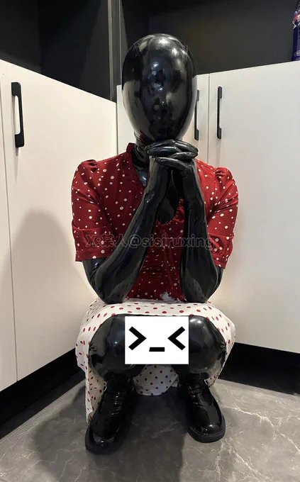 A shiny black latex doll figure squatting in a kitchen corner, wearing ordinary women's clothes, creating a surreal contrast between domestic life and fetish wear.