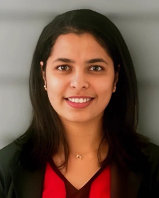 Professional headshot of a smiling individual wearing a black jacket over a red top, set against a blurred grey background.