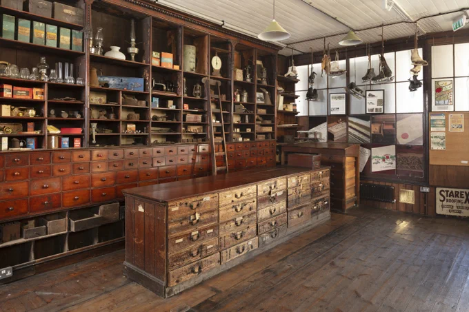 A photograph of a vintage general store interior with wooden cabinets filled with various items like lanterns, jars, and boxes.  Ice skates hang from the ceiling, and old advertisements and posters decorate the walls.