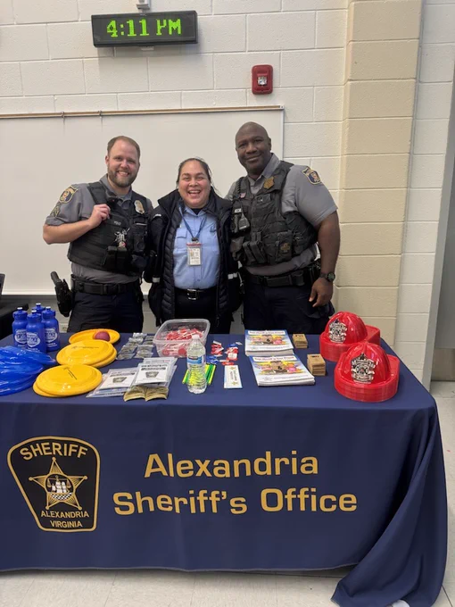 Three individuals, two in sheriff uniforms and one in a blue civilian outfit, stand smiling behind a table covered with promotional materials and labeled "Alexandria Sheriff's Office." The table also displays various giveaways including hats and brochures.