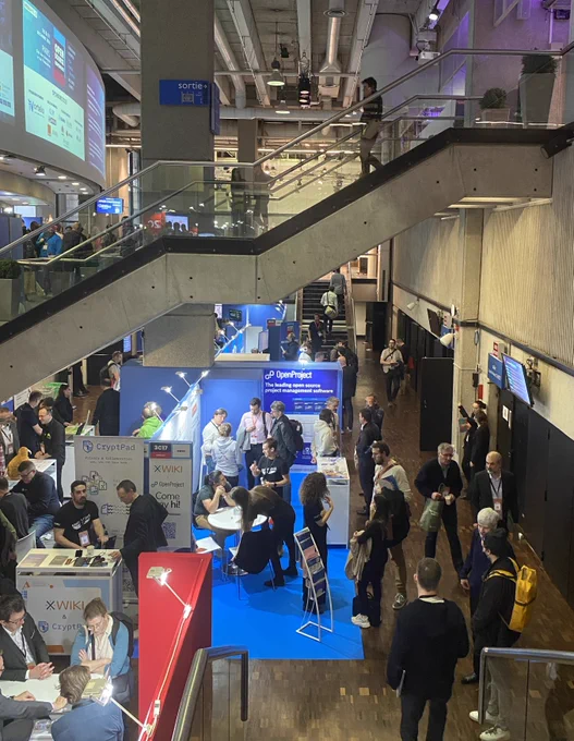 A wide view of the OSXP 2025 exhibition hall showing the OpenProject and XWiki booth among many attendees and stands.
