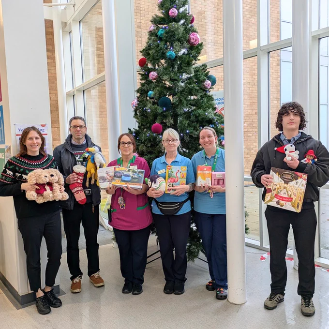 Six people stand in front of a Christmas tree, holding an assortment of cuddly toys, games and chocolates. The people are looking at the camera smiling.