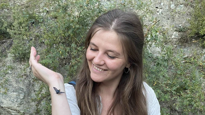 Charlotte Wright, a woman with long brown hair, smiles at a butterfly perched on her hand. She stands against a rocky backdrop with lush green foliage. 
