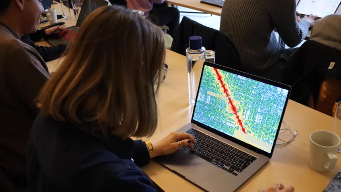A woman with light brown hair works on a laptop displaying a colorful, map-like graphic with red and yellow markings. She is seated at a wooden table with a water bottle and mug nearby. The setting is a focused, collaborative environment with others working on laptops.