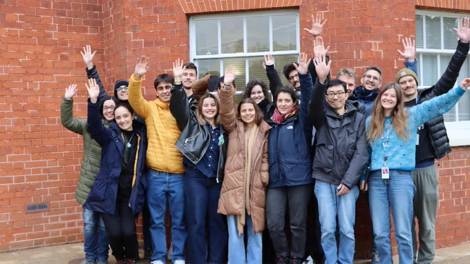 A group of 15 people stand smiling and waving in front of a brick building. They wear winter jackets, suggesting a cold day, and convey a cheerful, friendly atmosphere. It was a moment from the genome curation course run by Project Psyche at the Sanger Institute. Image credit: Carmen Denman Hume / Wellcome Sanger Institute.