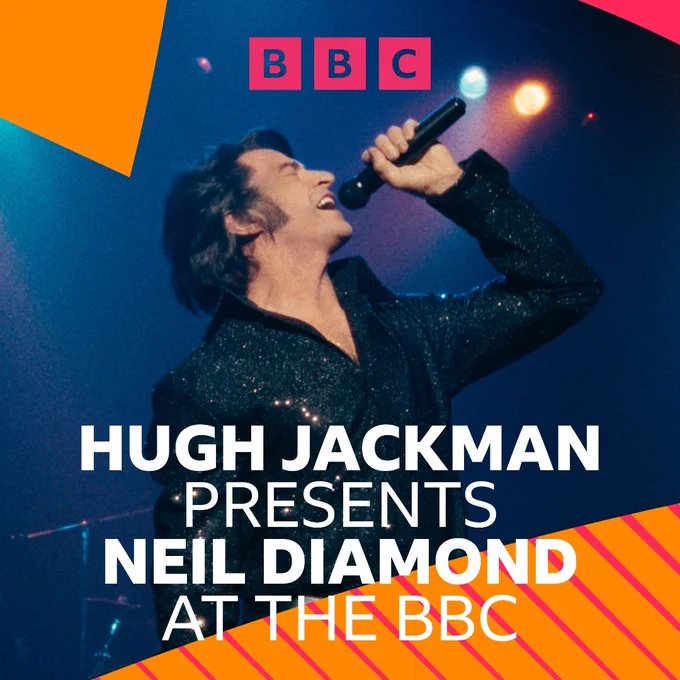 Neil Diamond wearing a black sparkly jacket singing into a handheld microphone under a spotlight