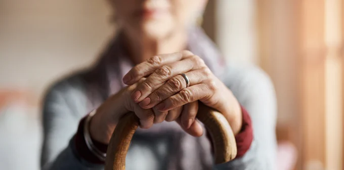 Close-up of an elderly person's clasped hands resting on the handle of a wooden cane