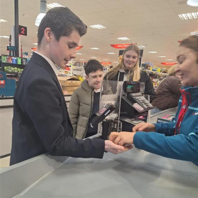 A cashier in a suit scanning a customer's item at a supermarket checkout counter with other shoppers waiting behind.
