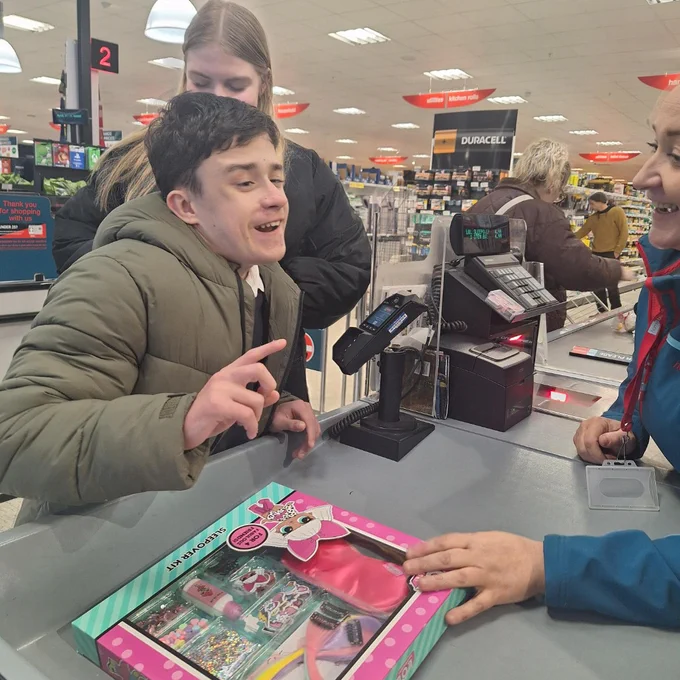 Child in a green jacket at a store checkout counter holding up a finger while a cashier smiles nearby.
