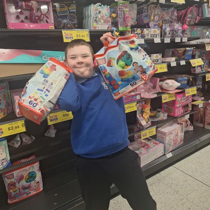 Child in blue sweater holding two large packs of colorful building blocks in a toy store aisle filled with various toys and price tags.