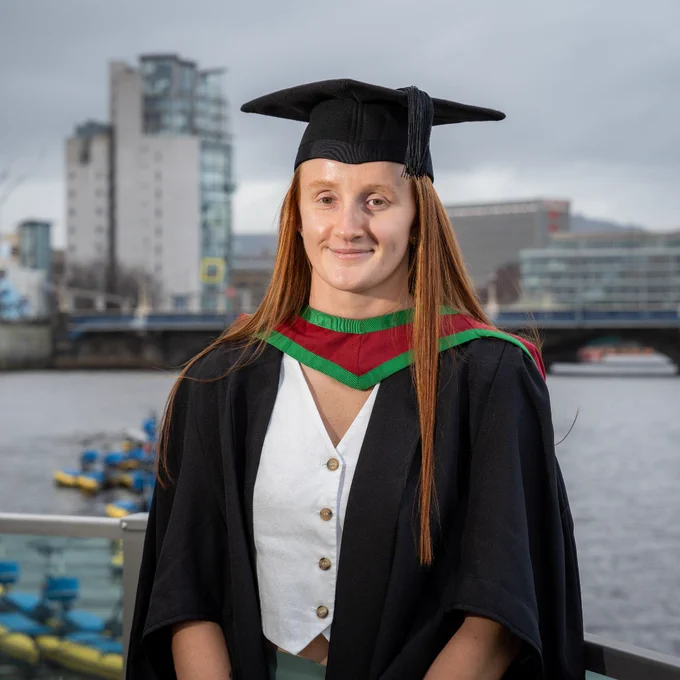 Graduate in black cap and gown with green and red trim standing by waterfront with city buildings in background.