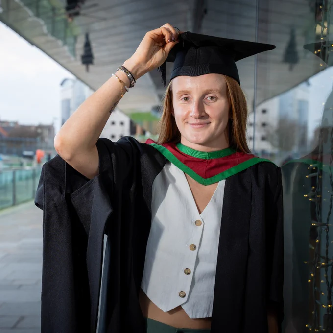 Graduate adjusting black mortarboard cap while wearing a gown with green and red hood outdoors.