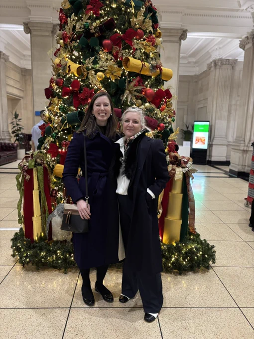 A photo of myself and Penelope. We are standing in front of a large Christmas tree, decorated red and gold. I am wearing a purple coat with a black bag. Penelope is wearing a black and white outfit, including black and white shoes, and a black fur gilet. We are both smiling. She has blonde hair and I have brown.