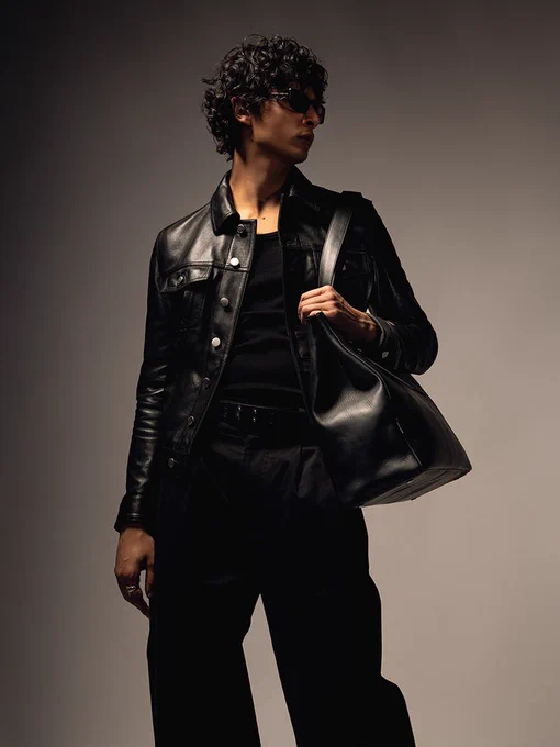 Studio portrait of a person dressed in all black—leather jacket, wide-leg trousers, and sunglasses—carrying a structured black leather shoulder bag.