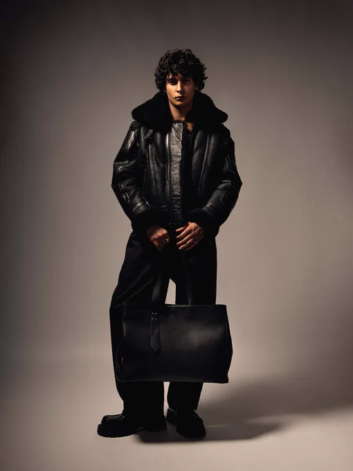 Studio portrait of a person in a black shearling jacket, chunky boots, and wide-leg trousers, carrying a structured black leather tote.