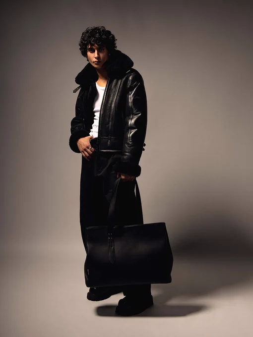 Studio portrait of a person in a black shearling jacket, white tank top, and loose black trousers, carrying an oversized black leather tote.