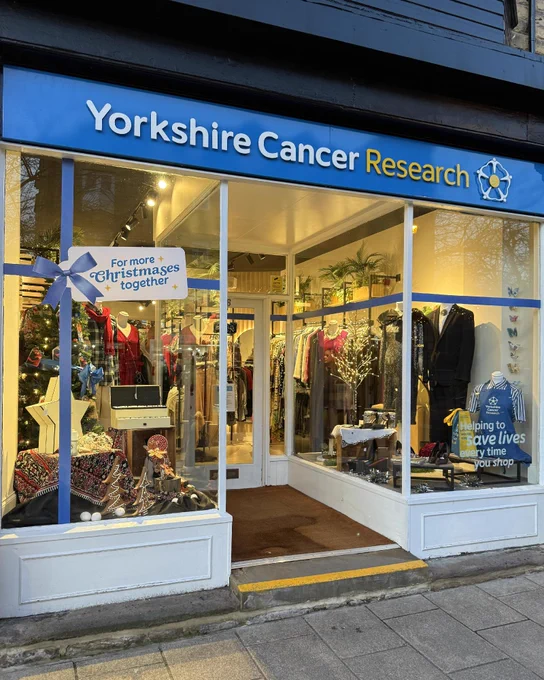 The front of the Yorkshire Cancer Research shop in Ilkley, decorated for Christmas with warm lights and clothing on display inside.