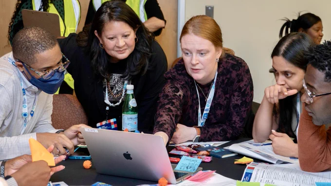 Group of people engaged in a discussion around a laptop at a table strewn with papers and office supplies.