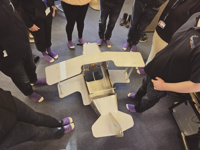 Group of people wearing matching purple striped socks standing around a metal frame resembling a small aircraft model on a carpeted floor.