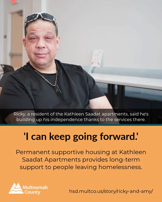 Person wearing sunglasses on his head and a black shirt sits in a room with tables, sharing his story about gaining independence through supportive housing.