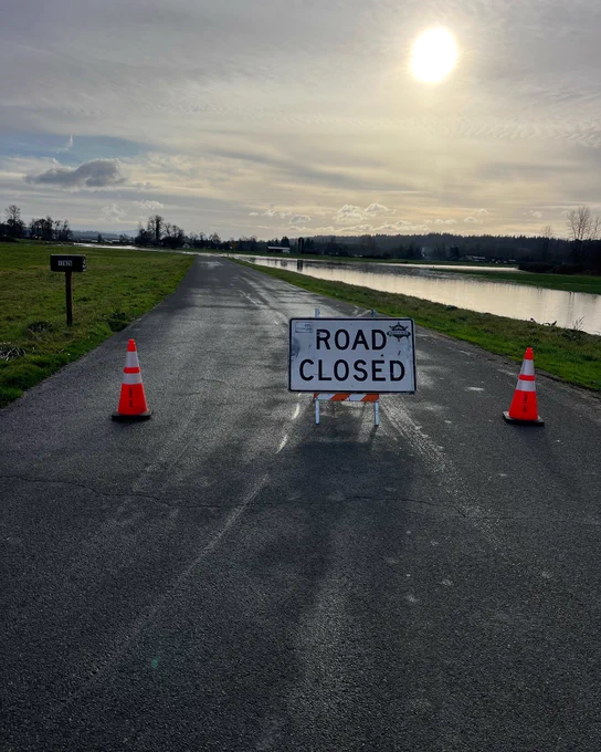 Road closure sign over flooded rural roadway. Pooling water can be seen in the background.