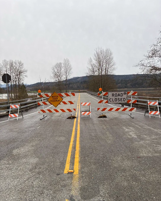 Large barricades block a roadway and have signage that reads "Water Over Roadway" and "Road Closed"