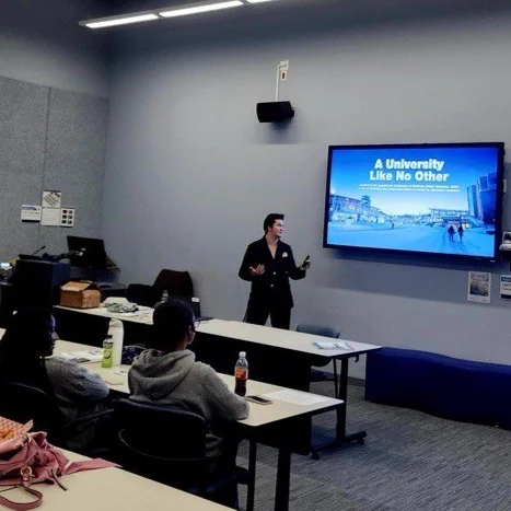 A person presents a slideshow titled "A University Like No Other" in a classroom setting with several attendees facing towards the screen.