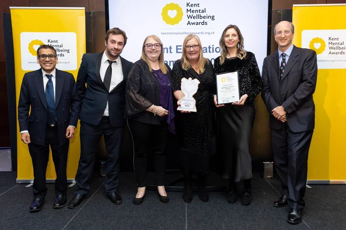 Six people are stood smiling in front of the Kent Mental Wellbeing Awards banners, two are holding the champions trophy and the award winner's certificate