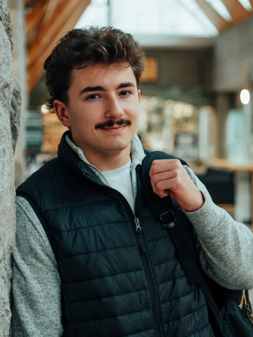 Person with a mustache wearing a navy puffer vest and grey shirt, smiling inside a warmly lit building.