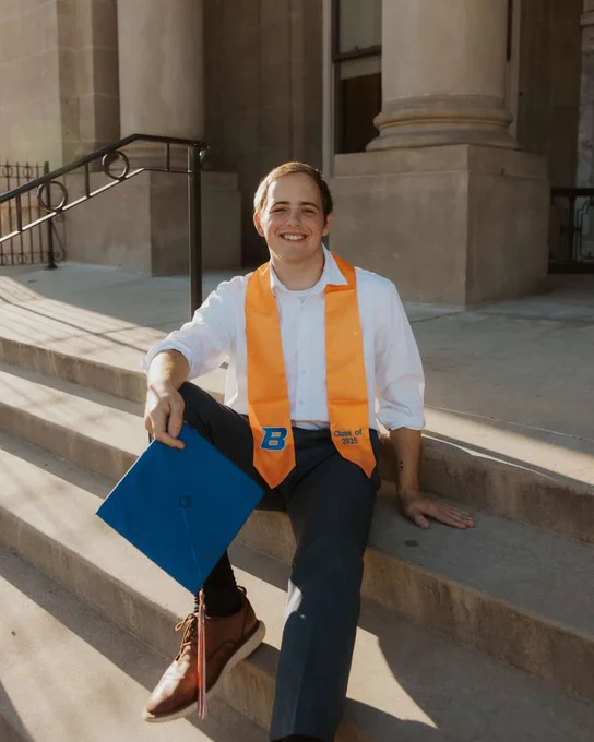 A Boise State student smiling wearing an undergraduate sash and holding a blue graduation cap, sitting on the stairs of a public building. 

