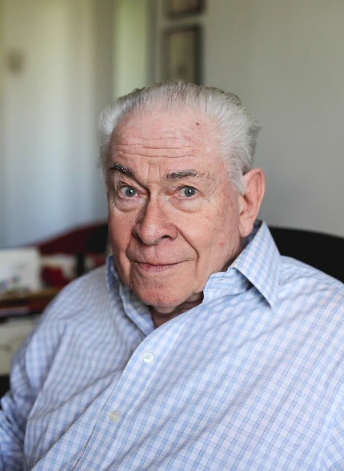 Scottish comedian and actor Stanley Baxter is pictured in a light blue checked shirt in a living room with white walls. He is gently smiling towards the camera and has combed back grey hair. 