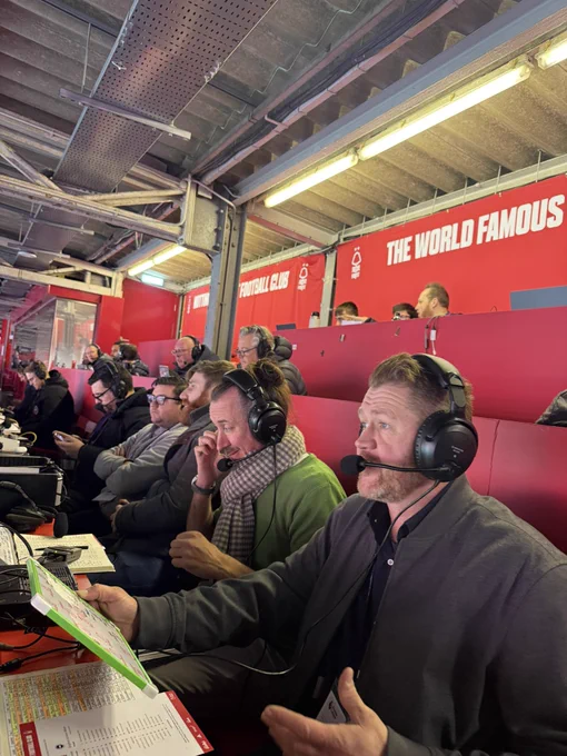 A busy press area with the main characters in this image sat facing to the left of the image. The commentators are wearing headset-microphones. They have paperwork and equipment in front of them. Winter clothing is being worn.