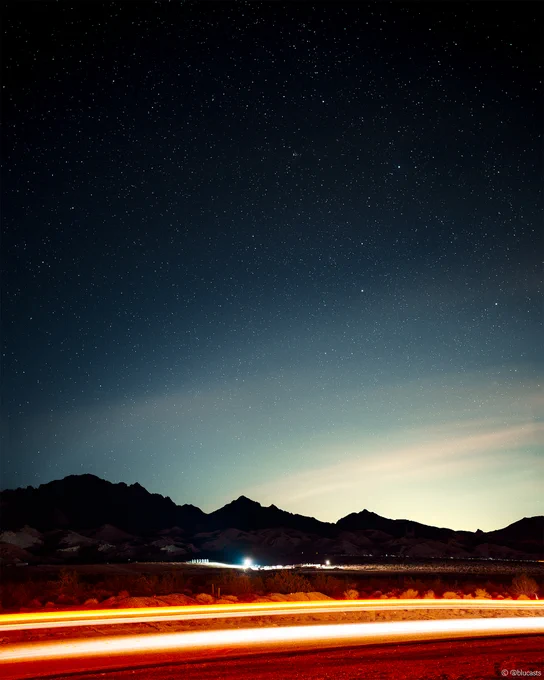 A long-exposure image of the night sky with mountains, the glow of a nearby city reflecting off of night time clouds, and an orange light beam from a passing car