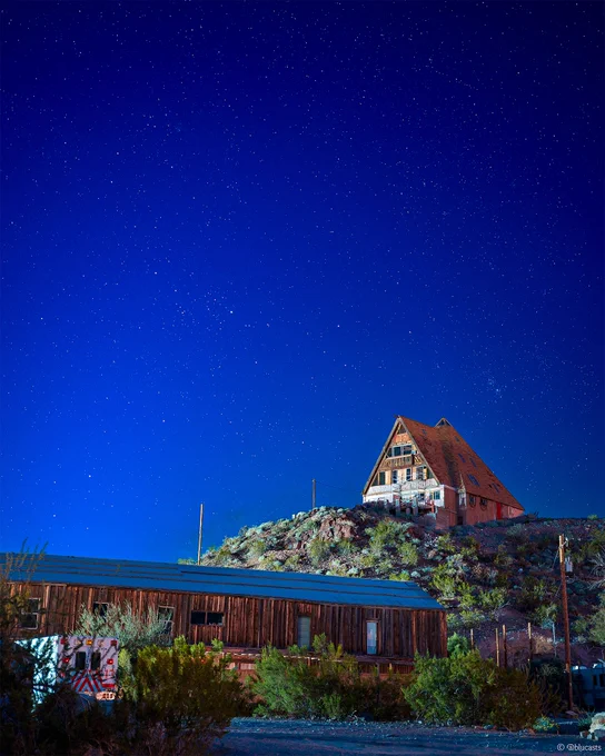 A near-dawn starscape with an incredibly vibrant blue hue centered by an A-Frame home on a hillside with green plants and an additional older wooden building and an ambulance in the foreground.