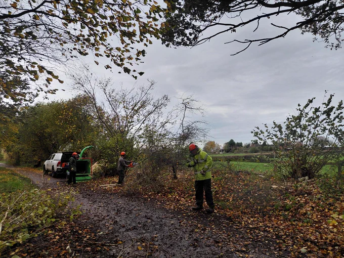 Two workers in high visibility jackets near a truck and a wood chipper in a rural setting with trees and an open field.