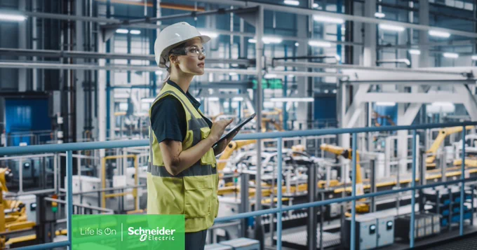 In einer Industrieumgebung ist eine Frau mit Schutzhelm und Schutzbrille zu sehen, die ein Tablet benutzt. Sie steht auf einer Plattform mit Blick auf Fabrikmaschinen. 