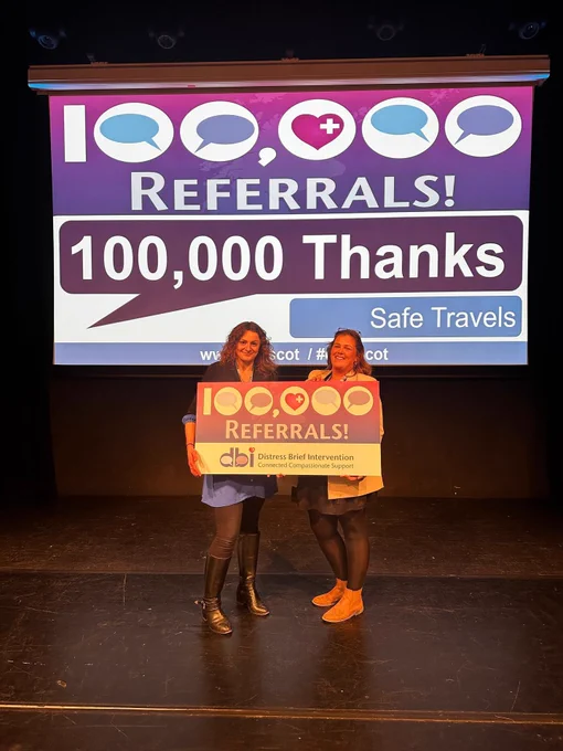 Two people holding a sign which says 100,000 referrals, Distress Brief Intervention. The screen in the background says the same.