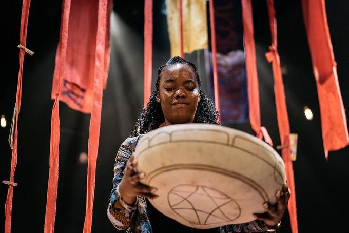 A Black, female actor performs on stage. Red strips of fabric hang behind her as part of the set, and she holds a large round bowl. Her eyes are closed. 