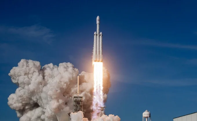 A powerful rocket launches into a clear blue sky, leaving behind a massive plume of white smoke and bright fiery exhaust. The scene captures the intensity and energy of the moment, with a nearby water tower visible in the background.