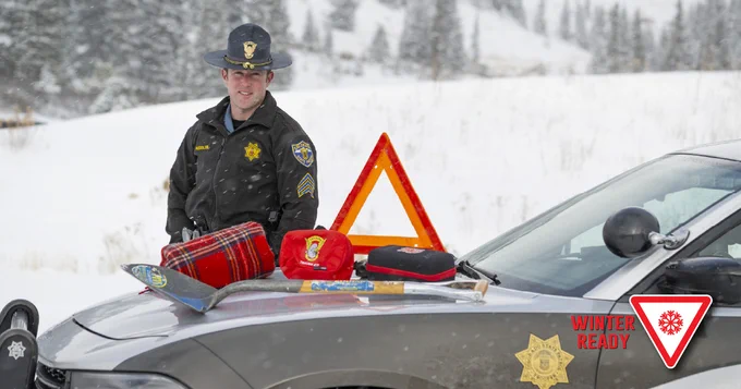 Trooper with patrol car, on car he has a blanket, medical bag, battery pack, shovel and orange triangle 