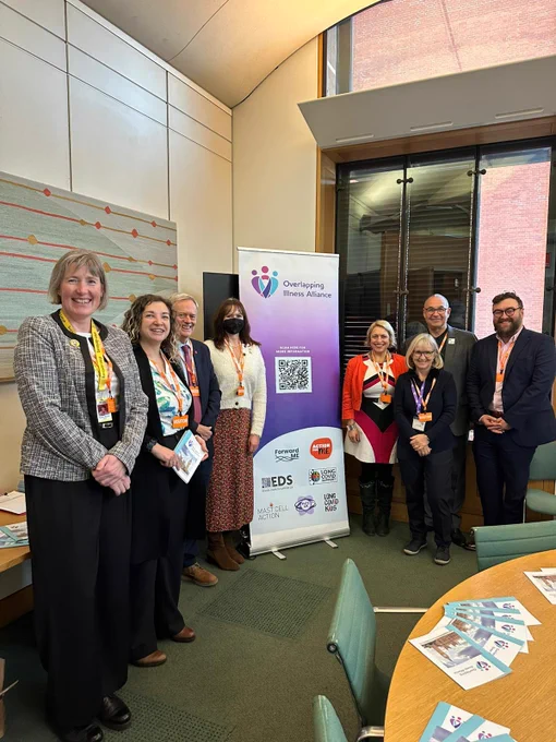 A group of eight people stand together in a meeting room beside a tall banner for the Overlapping Illness Alliance. The banner displays the Alliance logo and partner organisation logos. The group are smiling for the photo, and several are wearing visitor lanyards. A table in the foreground holds leaflets, and the room has large windows and modern wooden paneling.
