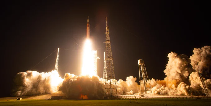A wide shot of Artemis I lifting off, as seen from the ground. The rocket, just left of center, has cleared the launchpad and is rising into the night sky, with a bright trail of light beneath it. Credit: NASA/Joel Kowsky