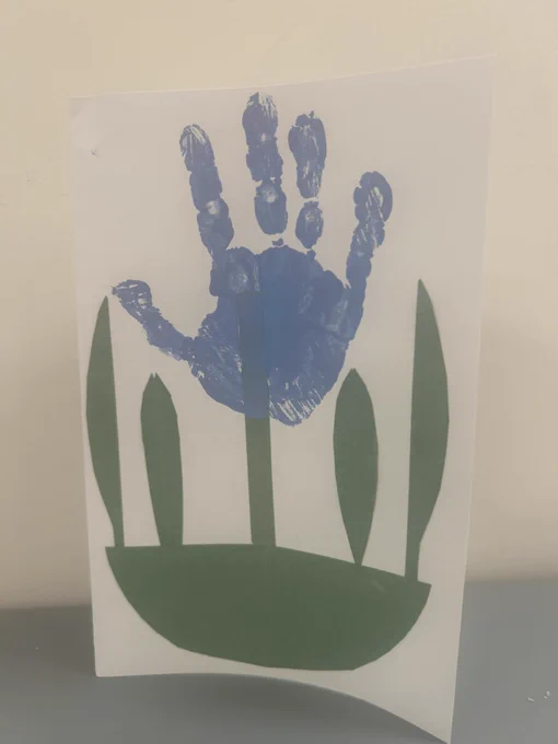 [Image 2 Description: Front of a handmade card. There is a blue child's handprint, on top of a green construction paper stem. There is a half-circle of green construction paper on the bottom, with green construction paper leaves, creating the image of a flower, with the handprint being the petals/flower. End image description.]